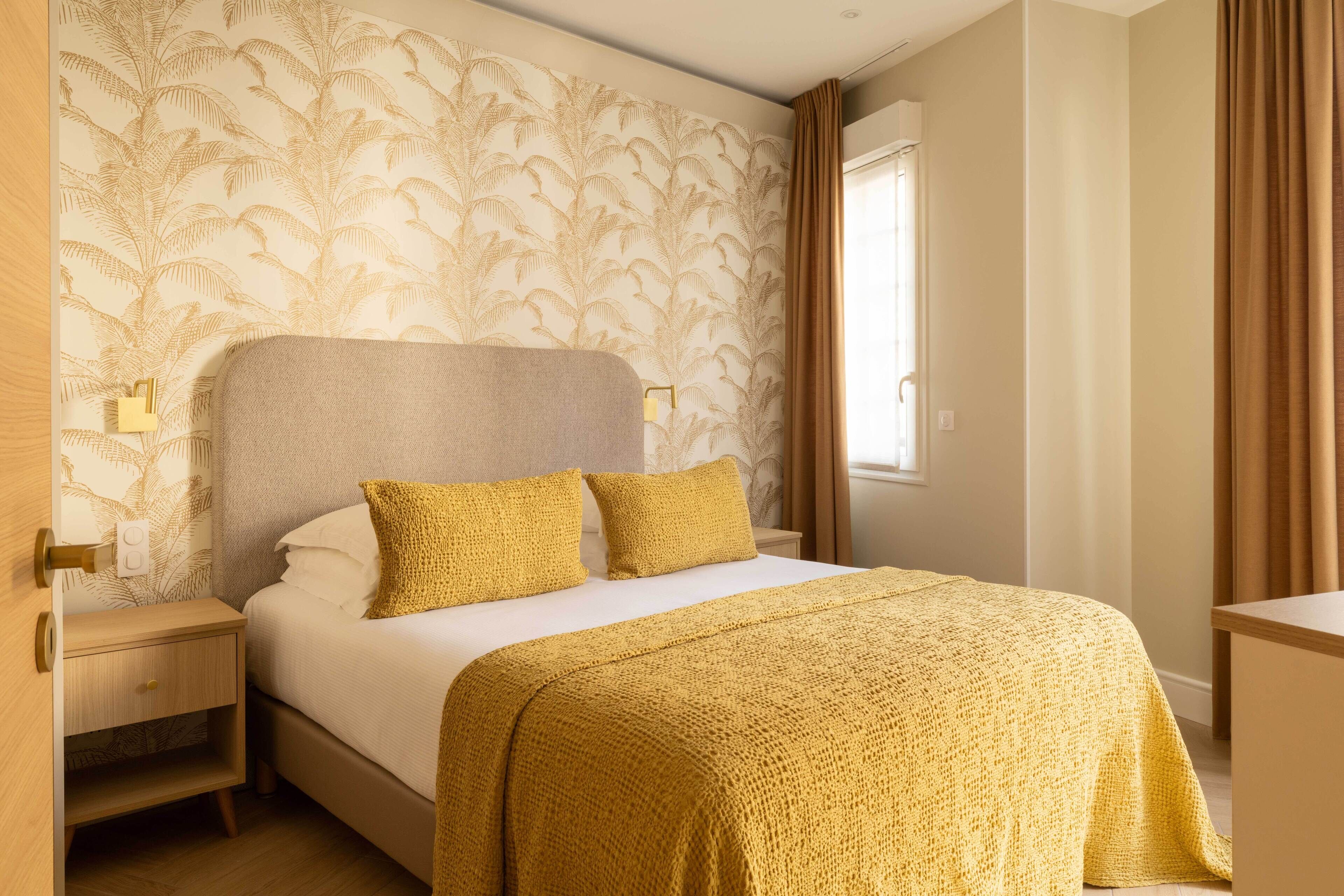 A neatly made bed with a textured blanket, a wooden nightstand with a drawer, and a wall with a leaf-patterned wallpaper.
