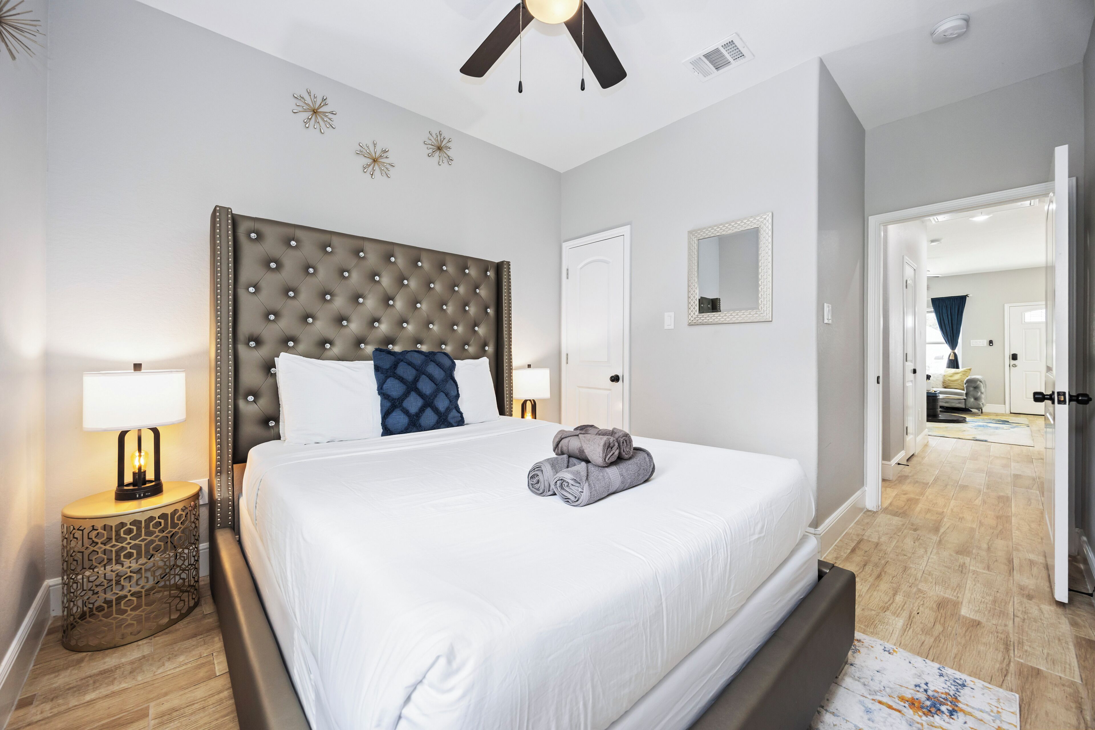 A hotel room with a large bed, a ceiling fan, a nightstand with a lamp, and a door leading to another room.