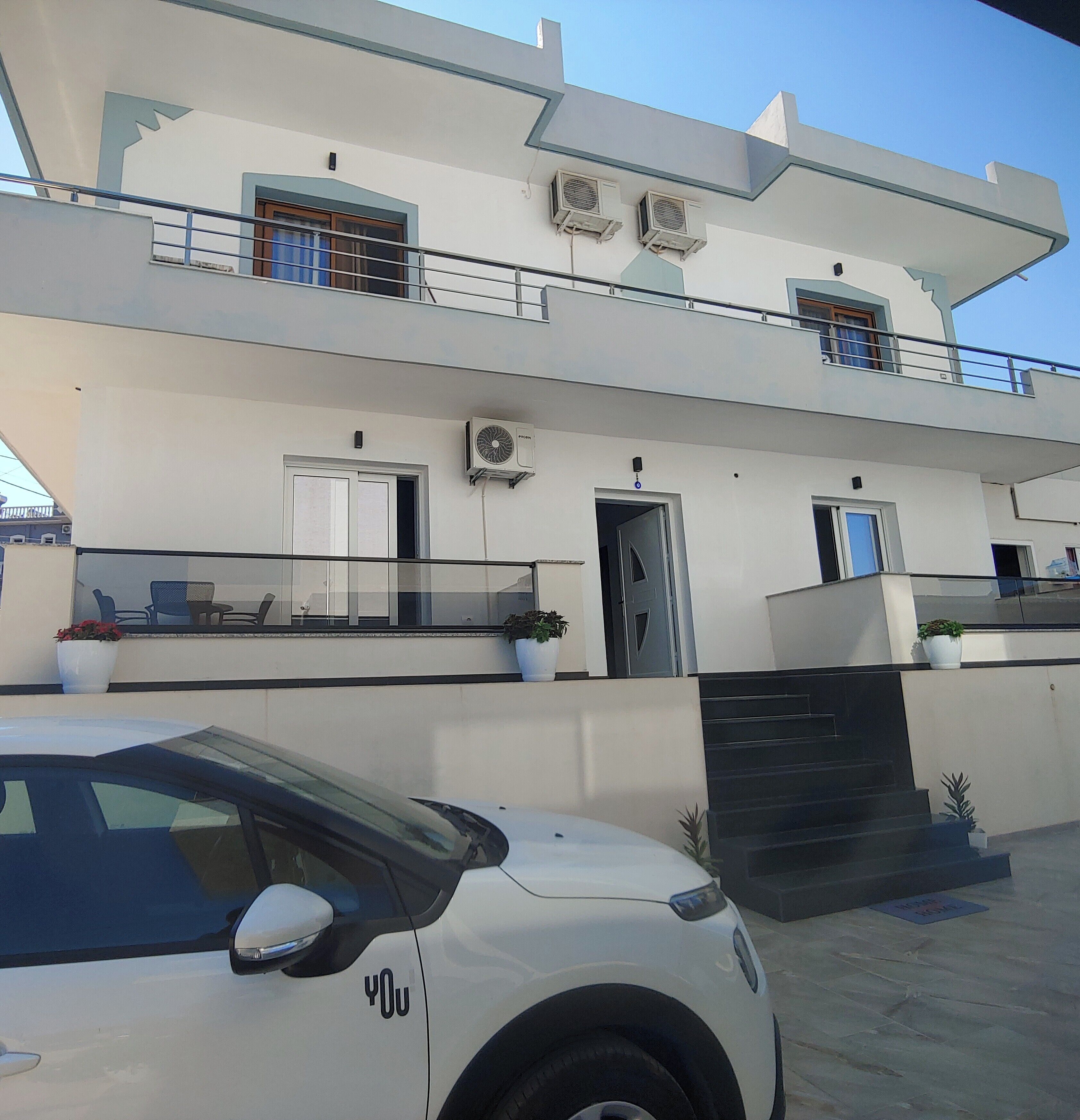 Un edificio moderno de dos pisos con un balcón, un coche estacionado frente a él y una escalera que conduce a la entrada.