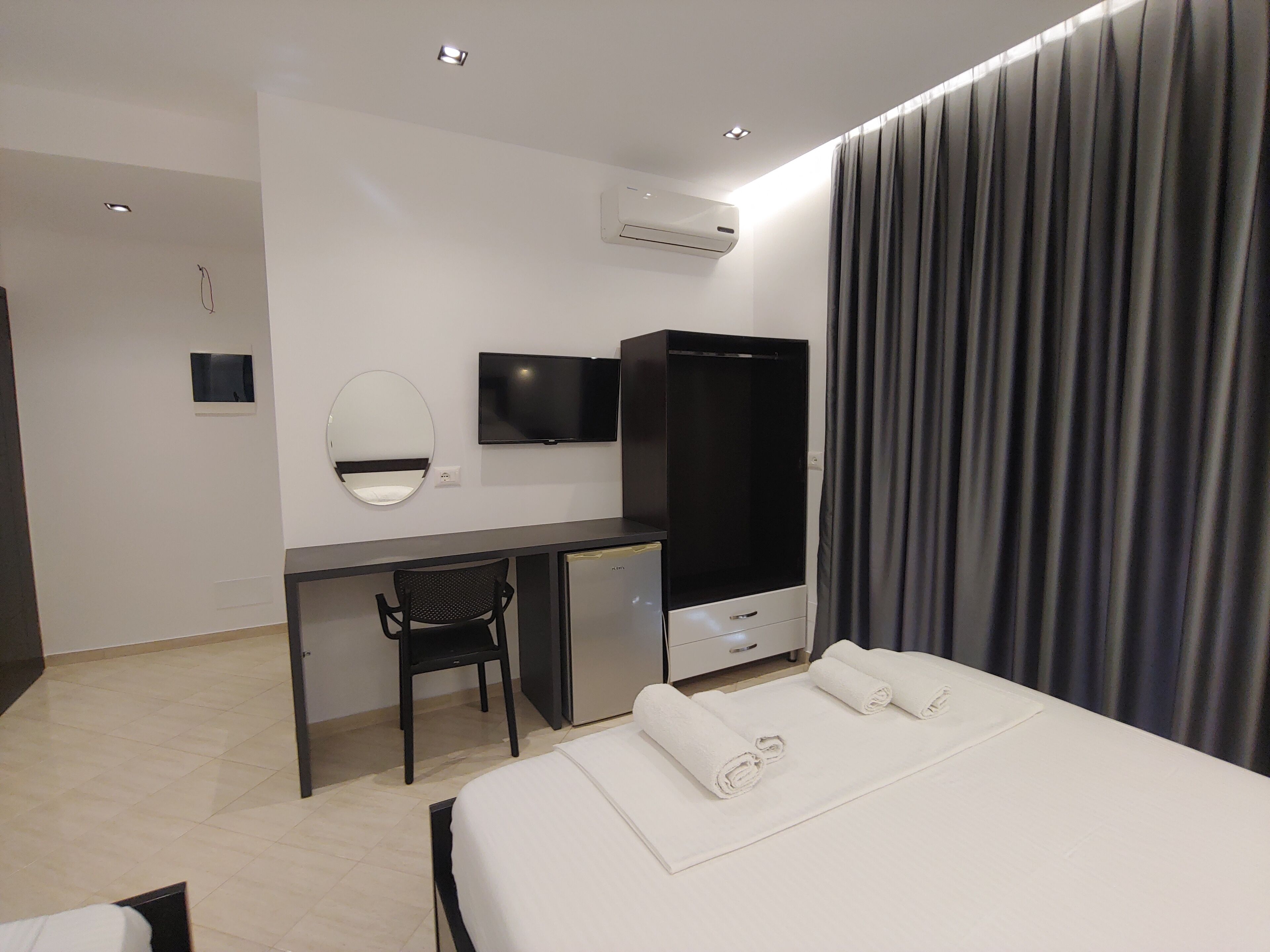 A modern hotel room with a bed, a desk with a chair, a TV, an air conditioner, and a large window with curtains.