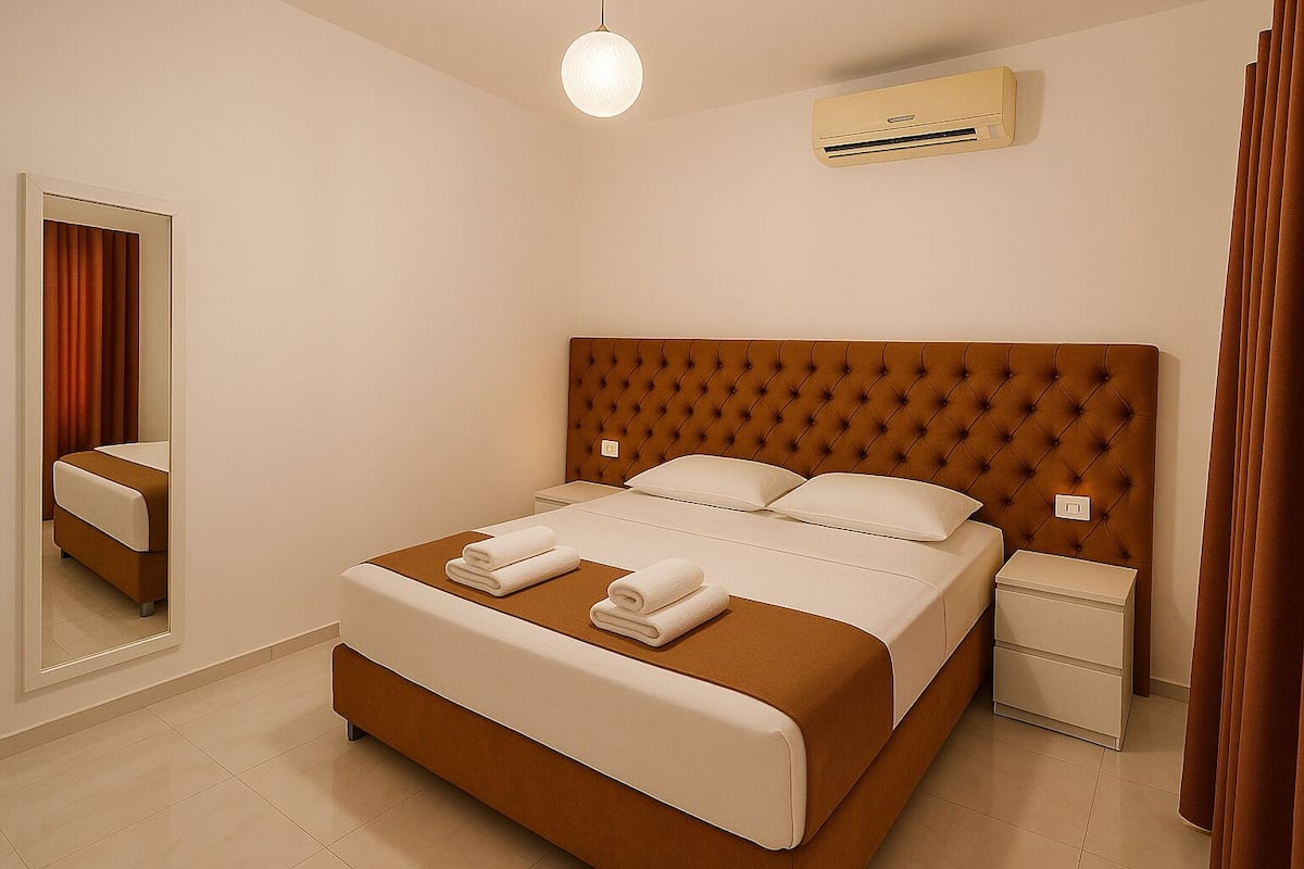 A neatly made bed with white linens and brown headboard, a bedside table, a wall-mounted air conditioner, and a mirror reflecting the room.