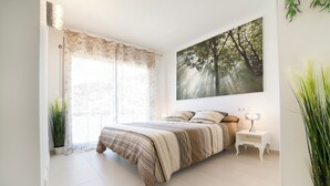 A bedroom with a large bed, a nightstand, a wall-mounted lamp, and a forest-themed painting. - Duplex in Sitges with sea views, parking, terrace, balcony (Sitges)