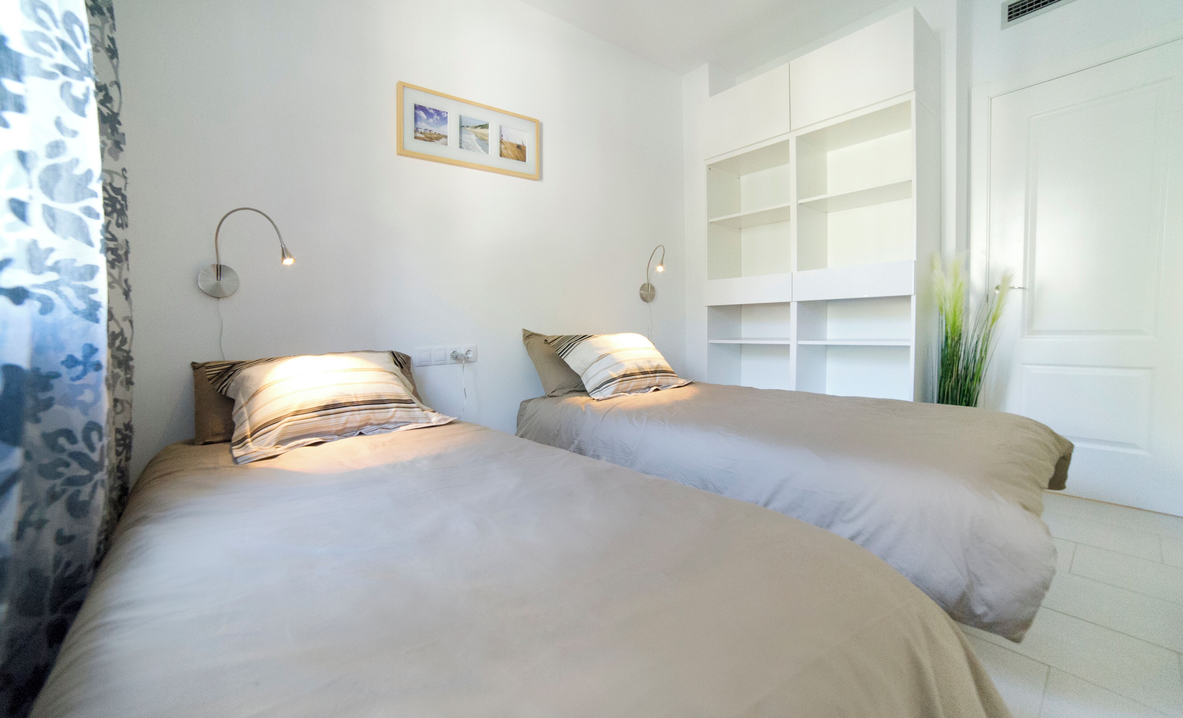 A bedroom with two beds, a wall-mounted shelf unit, and a window with blinds.