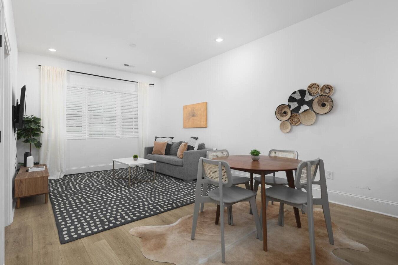 A modern living room with a grey sofa, a white coffee table, and a round wooden dining table with grey chairs. There is a black and white patterned rug, a wooden floor, and a wall-mounted woven art piece.