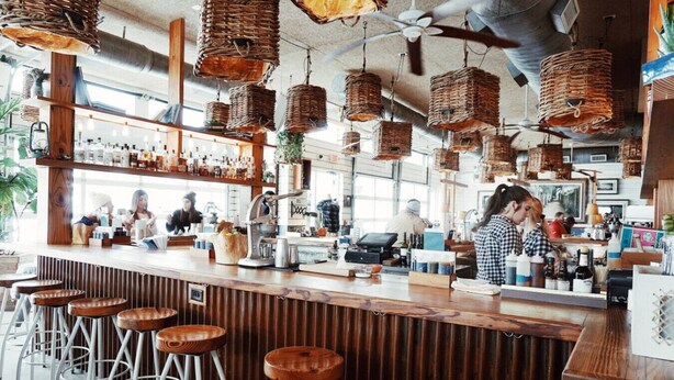 A bar with wooden bar stools, hanging wicker baskets, and a counter with baristas preparing drinks.