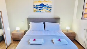 Una cama bien hecha con sábanas blancas y dos toallas con flores moradas, un cuadro de montañas en la pared y lámparas de noche sobre mesitas de madera.