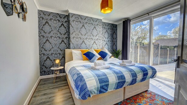 A bedroom with a large bed, a bedside table with a lamp, patterned wallpaper, and a view of the outdoors. - 4 BedRoom House | w/ FREE Wi-fi | Parking (West Midlands)