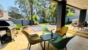 An outdoor patio with a grill, green and yellow chairs, a table with a vase, and a view of the backyard.