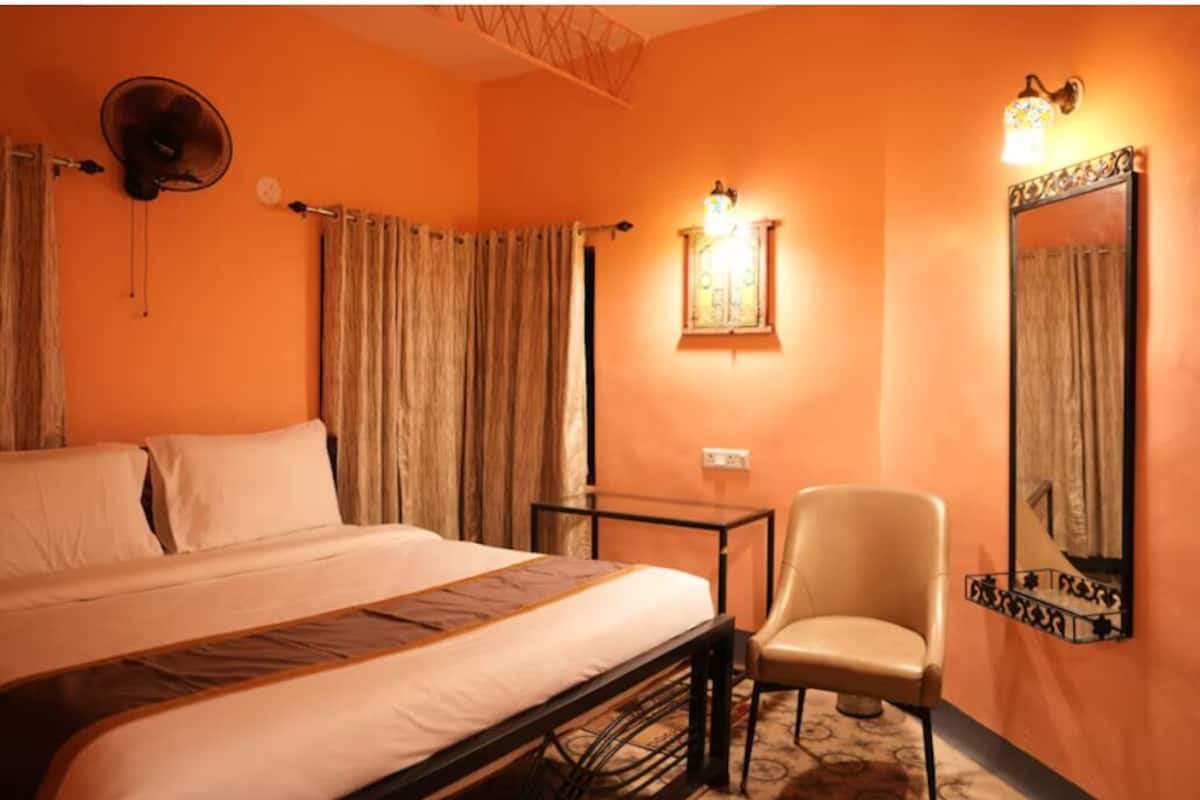 Une chambre d’hôtel comprenant un lit, une chaise, une table, des rideaux, un ventilateur fixé au mur et un luminaire fixé au mur.