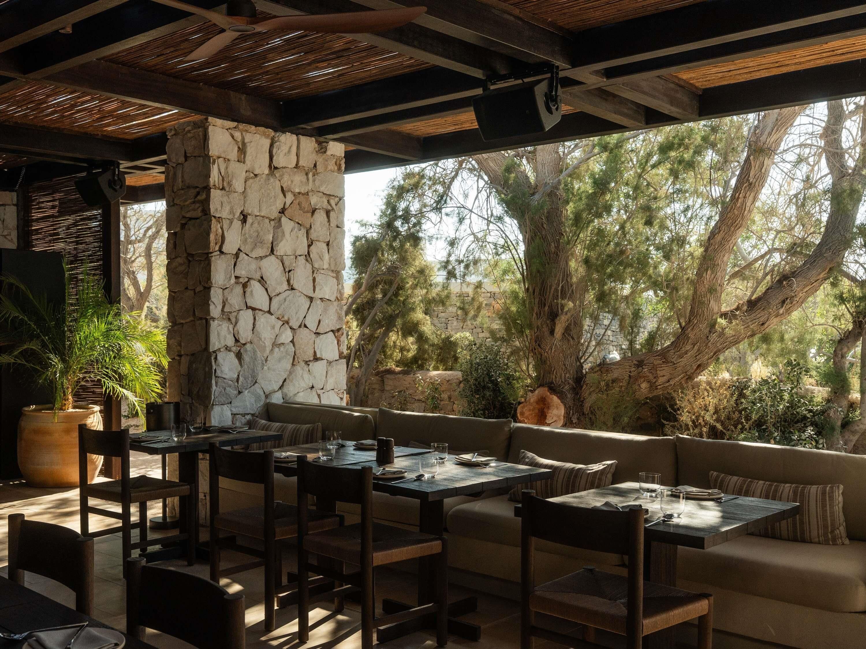 An outdoor seating area with stone walls, wooden furniture, and a view of trees and sky.