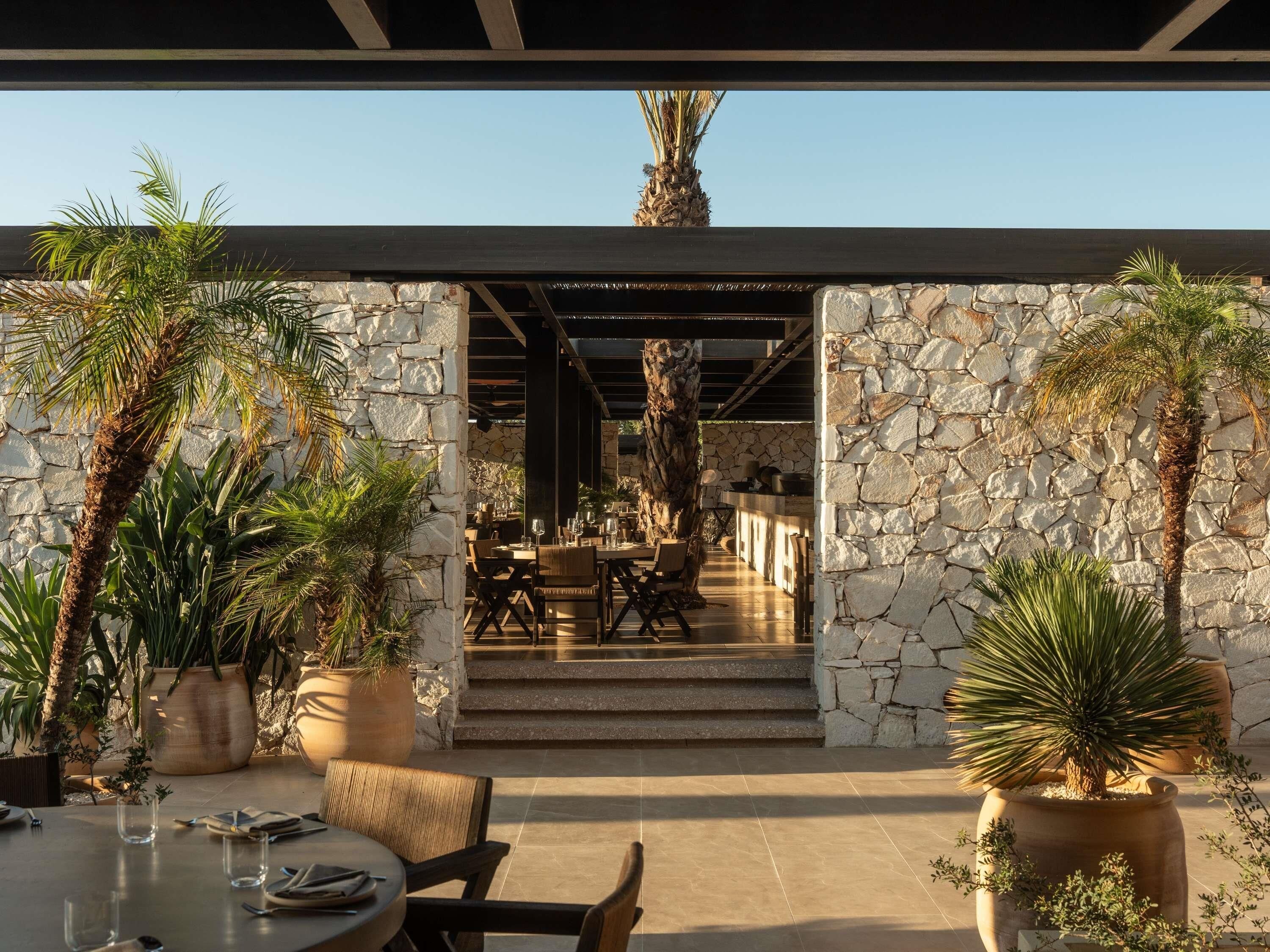 An outdoor dining area with stone walls, palm trees, and a view into a covered dining space.