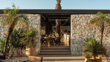 An outdoor dining area with stone walls, palm trees, and a view into a covered dining space.