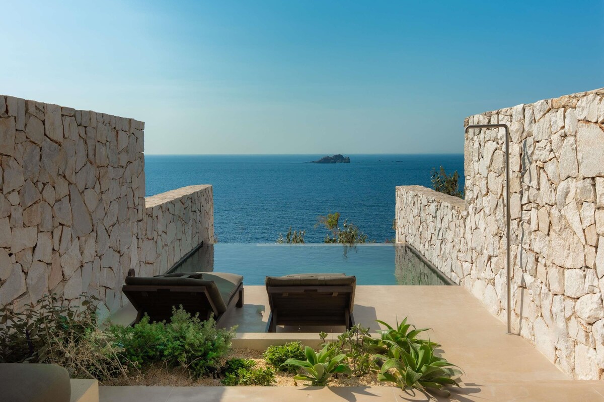 An infinity pool with stone walls and lounge chairs, overlooking a calm sea and a small island.