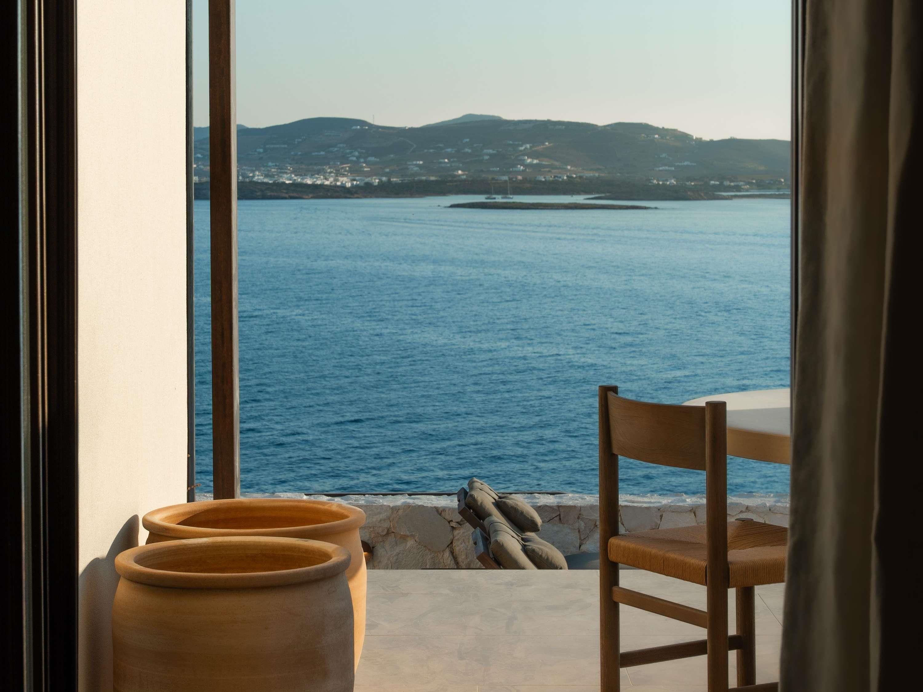 Vue depuis une pièce dotée d’une grande fenêtre donnant sur une étendue d’eau et sur des collines lointaines. On y aperçoit une chaise et une table sur laquelle sont posés un vase et une plante.