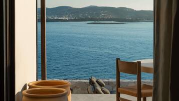 Vue depuis une pièce dotée d’une grande fenêtre donnant sur une étendue d’eau et sur des collines lointaines. On y aperçoit une chaise et une table sur laquelle sont posés un vase et une plante.