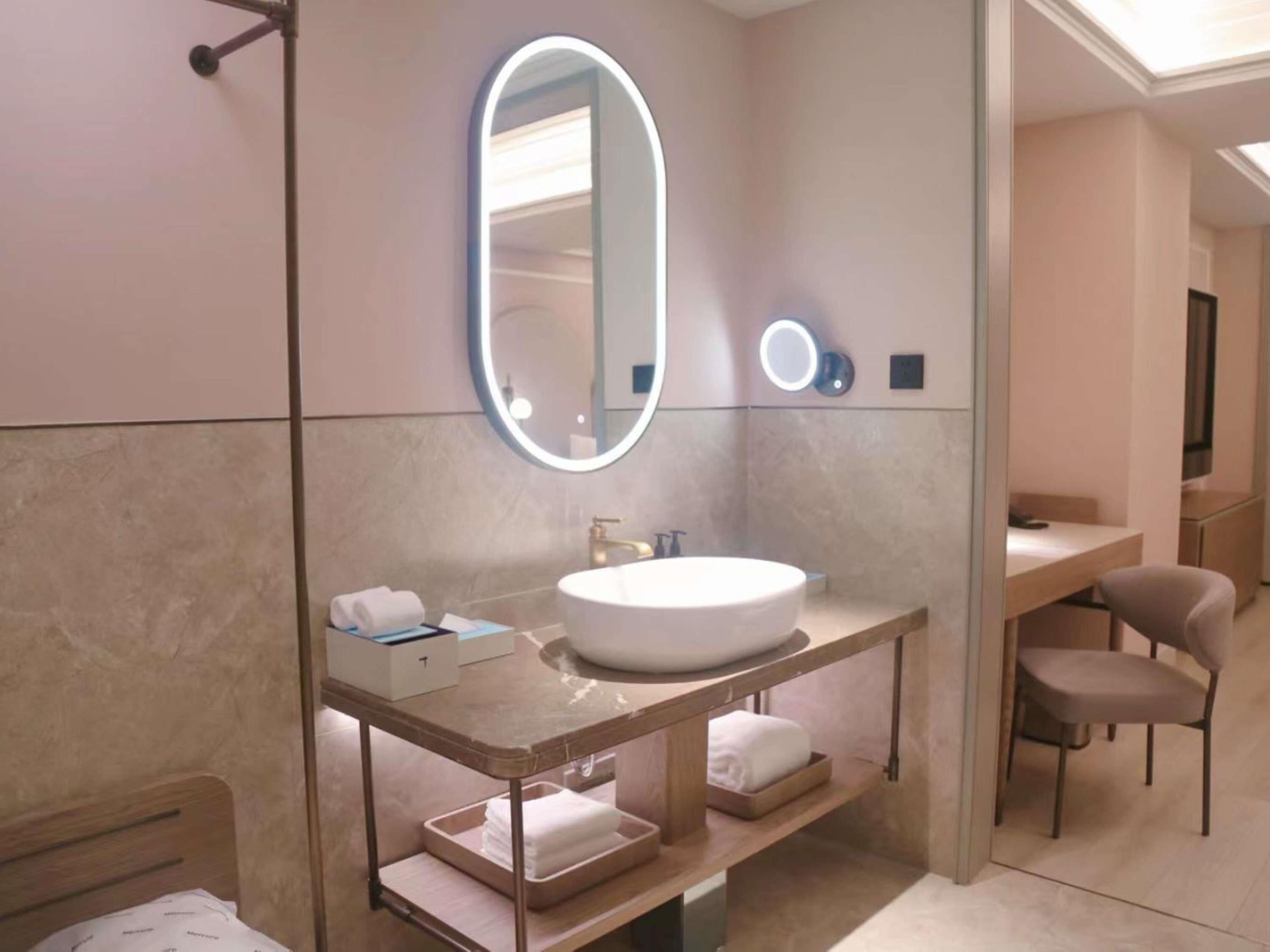 Un baño moderno con un espejo ovalado grande, un lavabo blanco y un mueble de tocador con estantes para toallas.