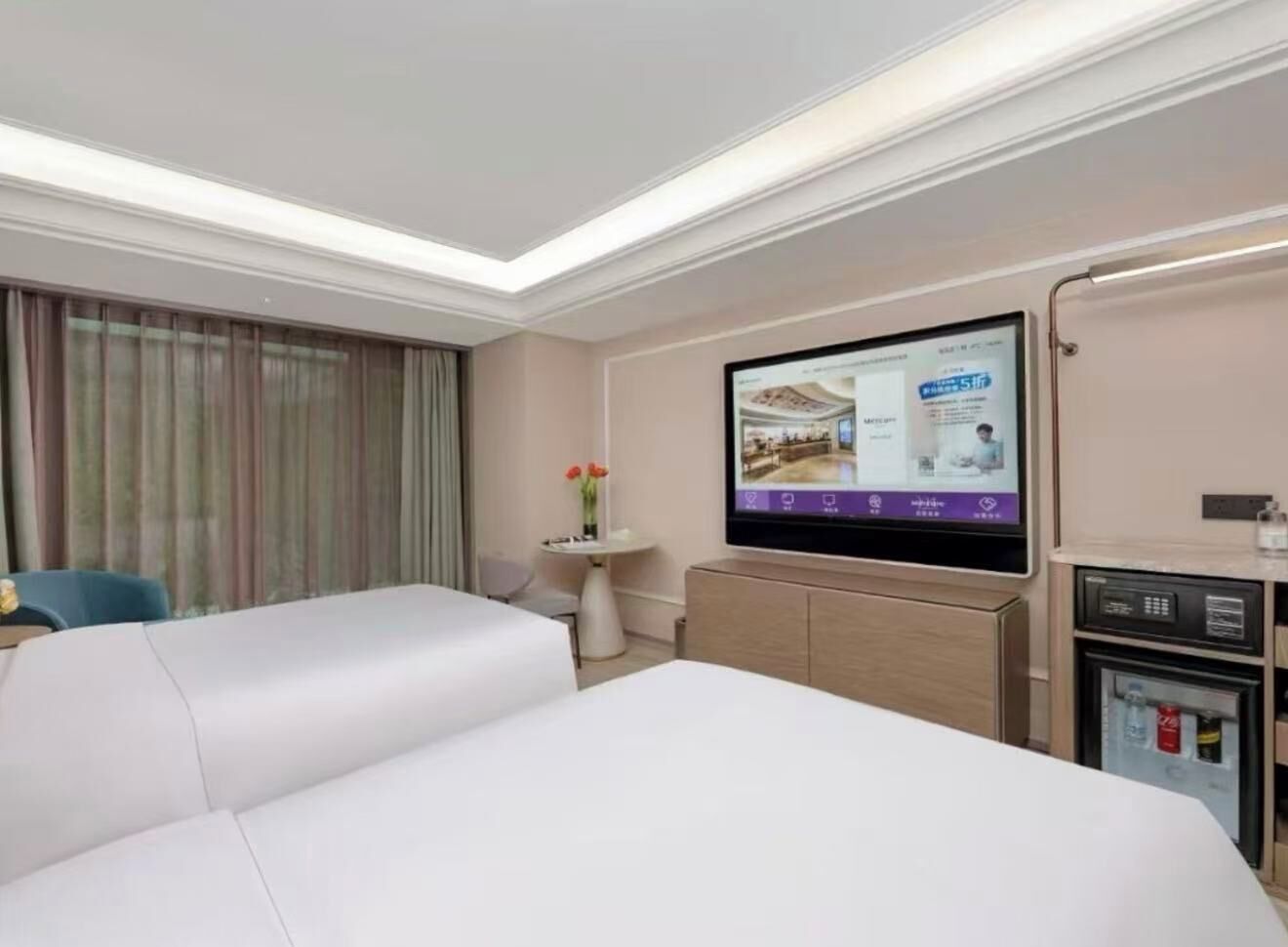 A hotel room with a large bed, a television, a small table with a vase, and a minibar.