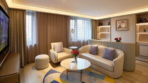 Un soggiorno moderno con un divano, una poltrona, un poggiapiedi e un tavolino da caffè. Si nota una libreria con libri e oggetti decorativi, e una televisione montata a parete.