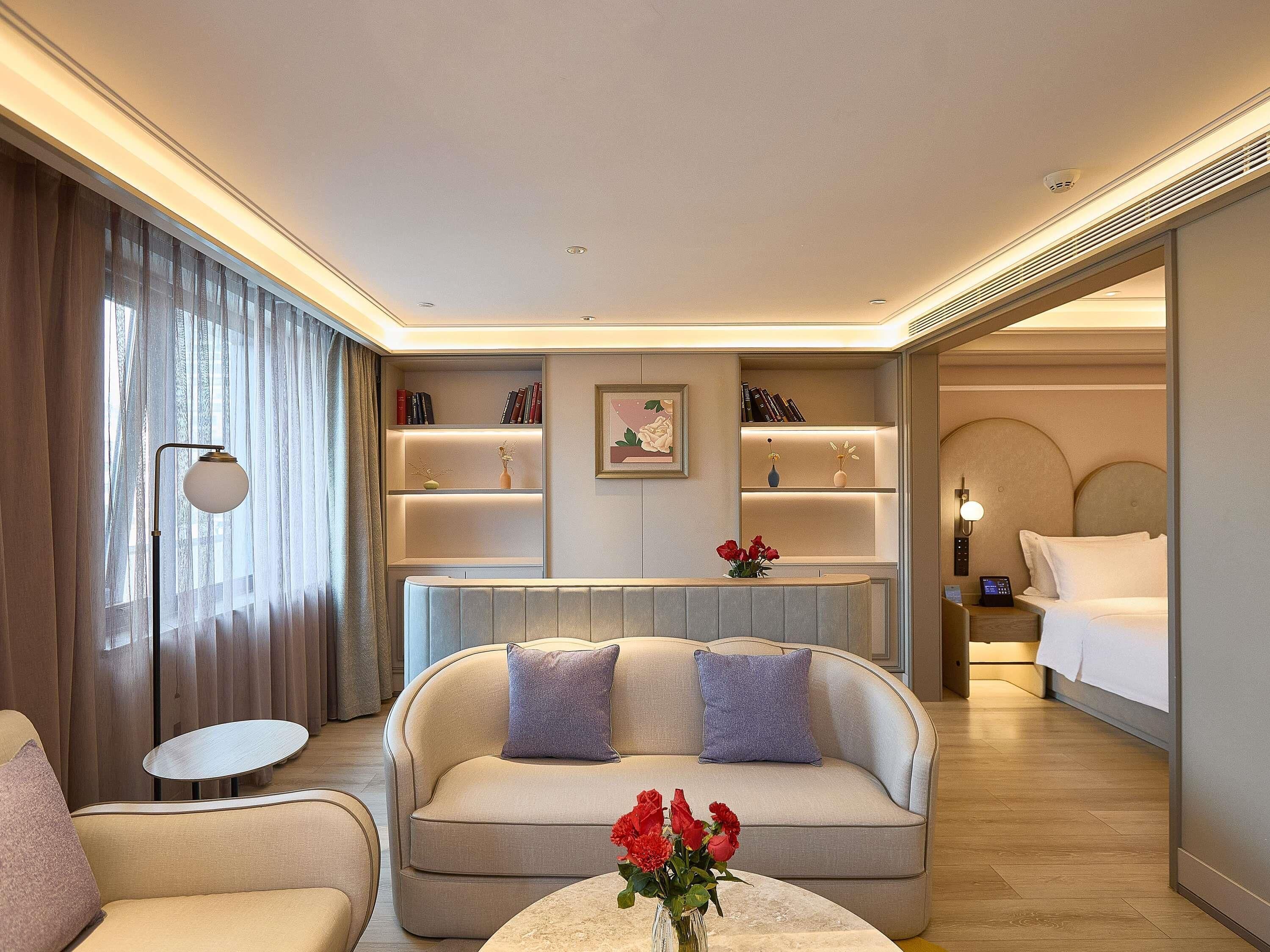 A modern hotel room with a sofa, a small round table, a bedside table with a lamp, a bed, and a wall-mounted shelf with books and decorative items.