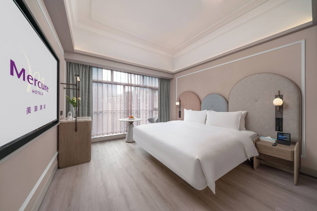 A hotel room with a large bed, bedside tables, a small table with a vase, and a large window with curtains.