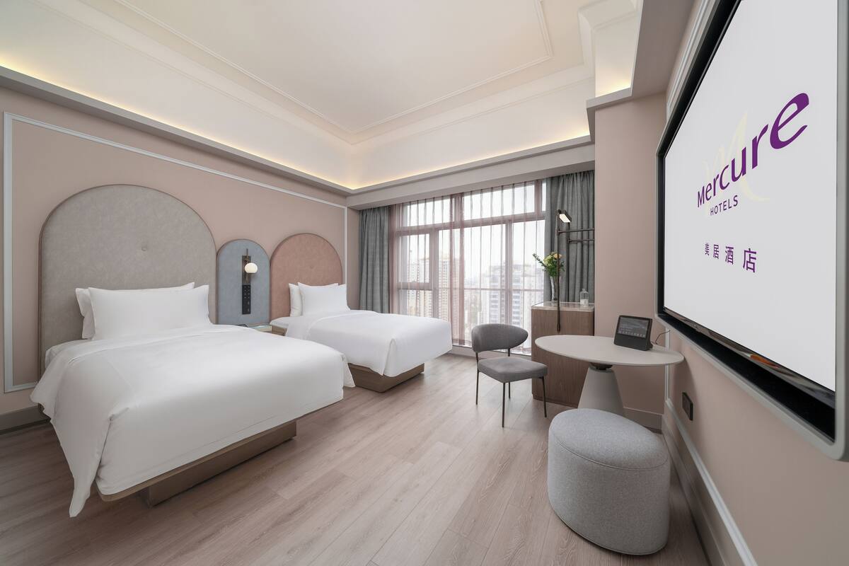 A hotel room with two beds, a small table, a chair, and a large window.