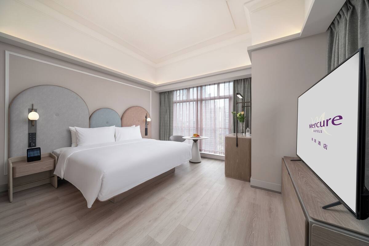 A hotel room with a large bed, bedside tables, a TV, and a view of the city through large windows.