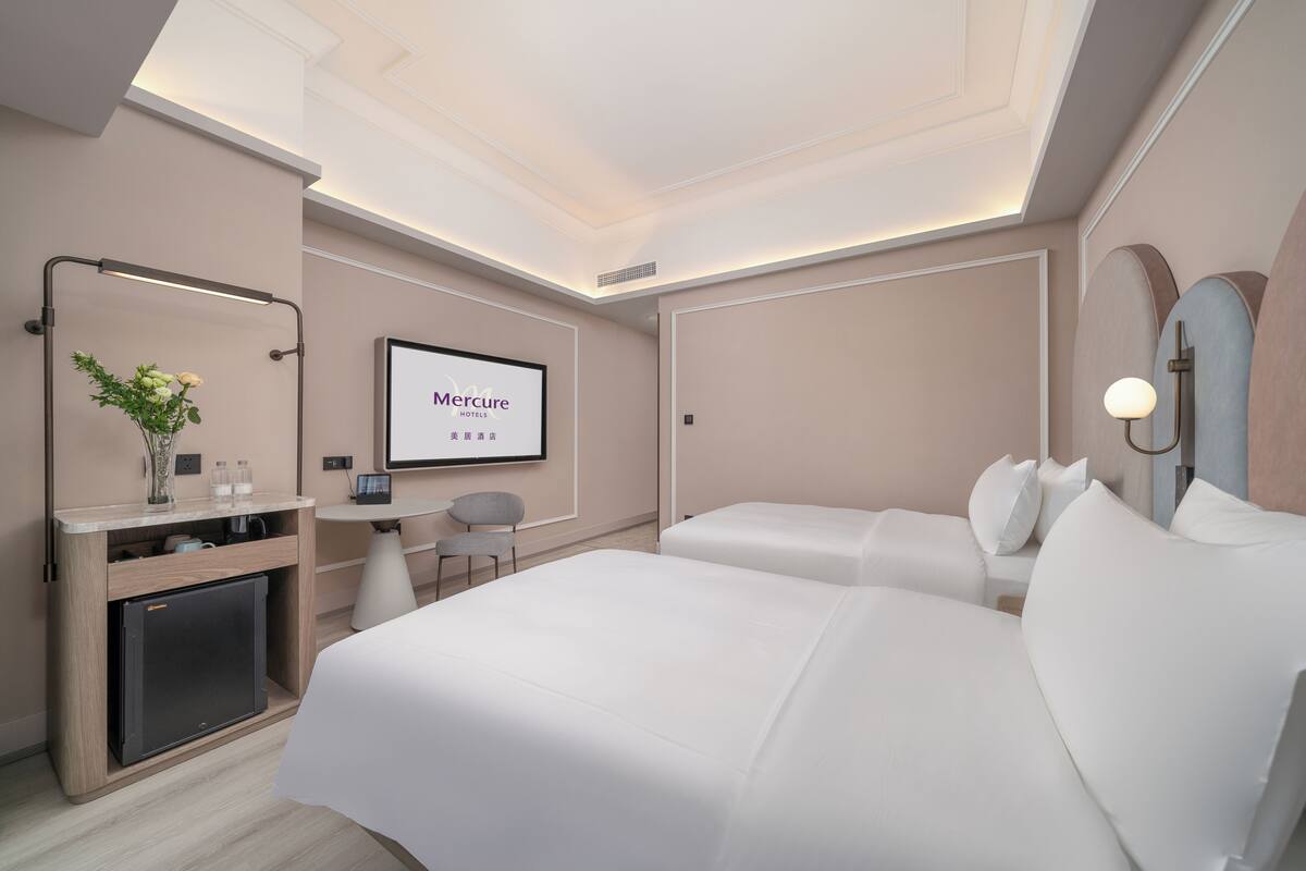 A hotel room with a bed, a desk with a computer, a small fridge, a TV, and a wall-mounted mirror.
