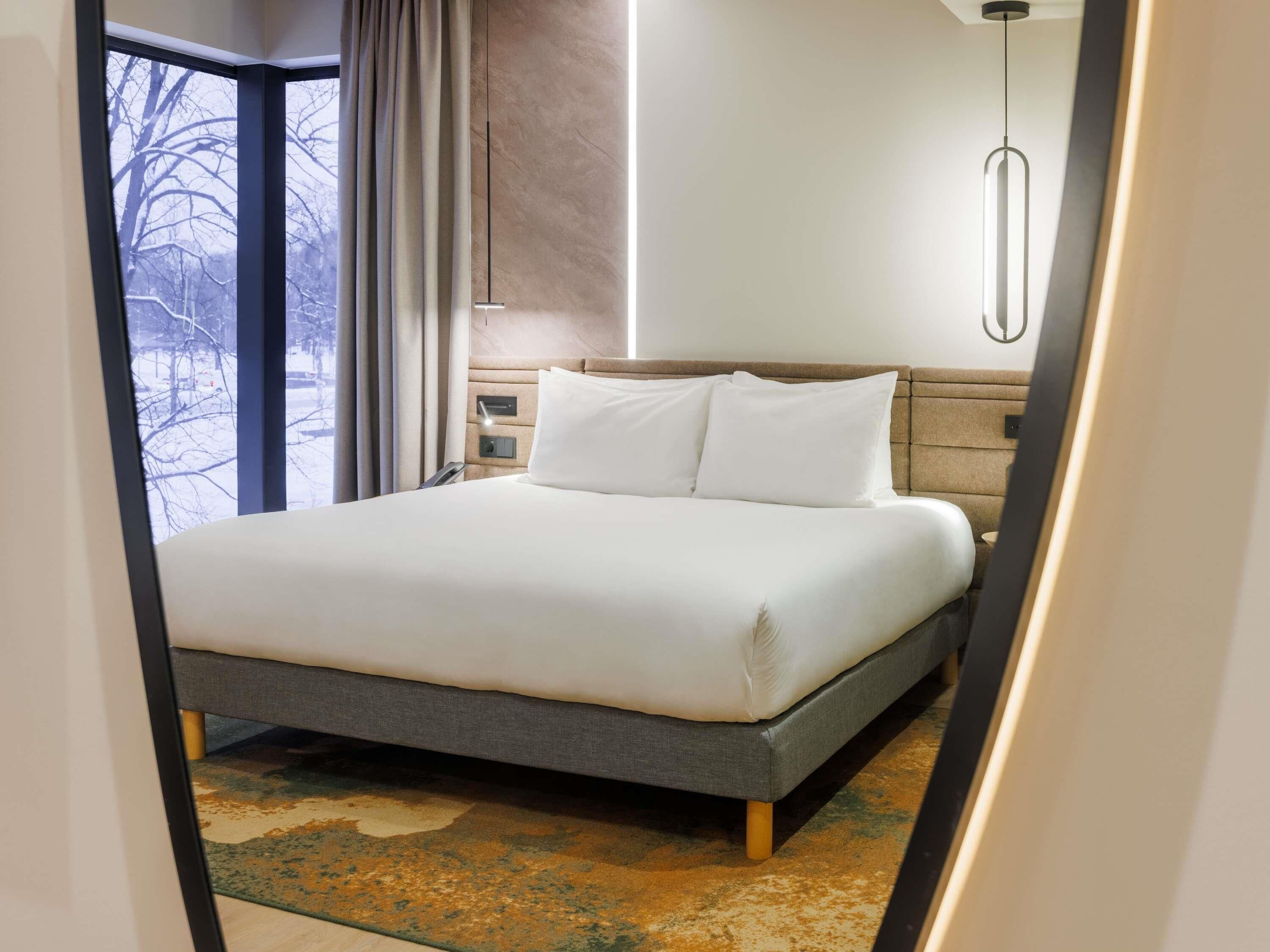 A modern hotel room with a large bed, a nightstand, a bedside table, a wall-mounted light, and a view of a snowy landscape through a window.