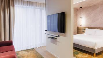 Une chambre d’hôtel avec un lit, un téléviseur fixé au mur, un fauteuil rouge et une grande fenêtre avec des rideaux.