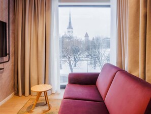 A room with a red sofa, a small round table, and a window with a view of a church and trees.
