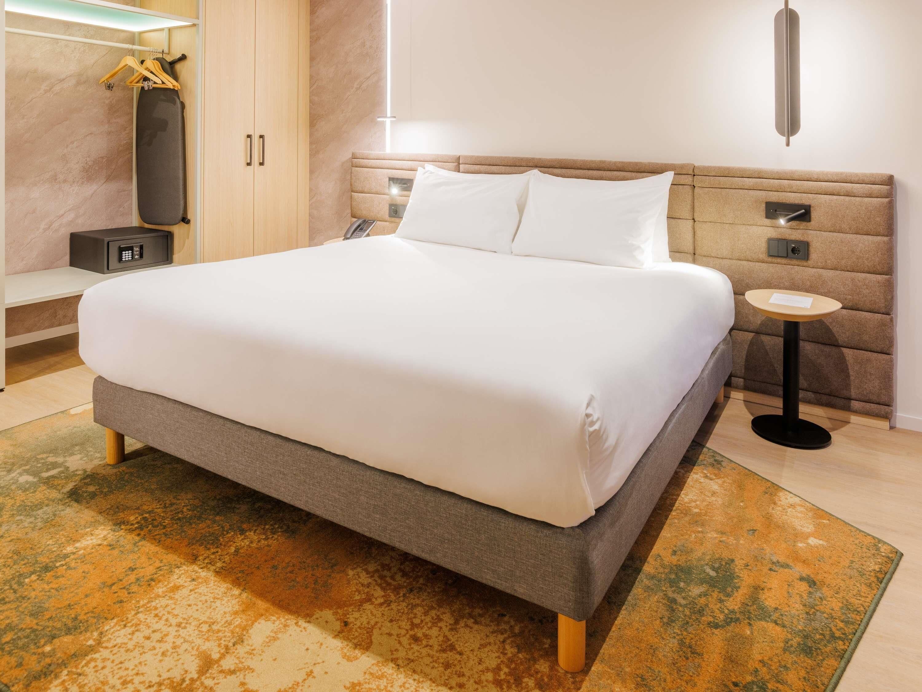 A modern hotel room with a large bed, a bedside table, a wardrobe, and a small table with a lamp.