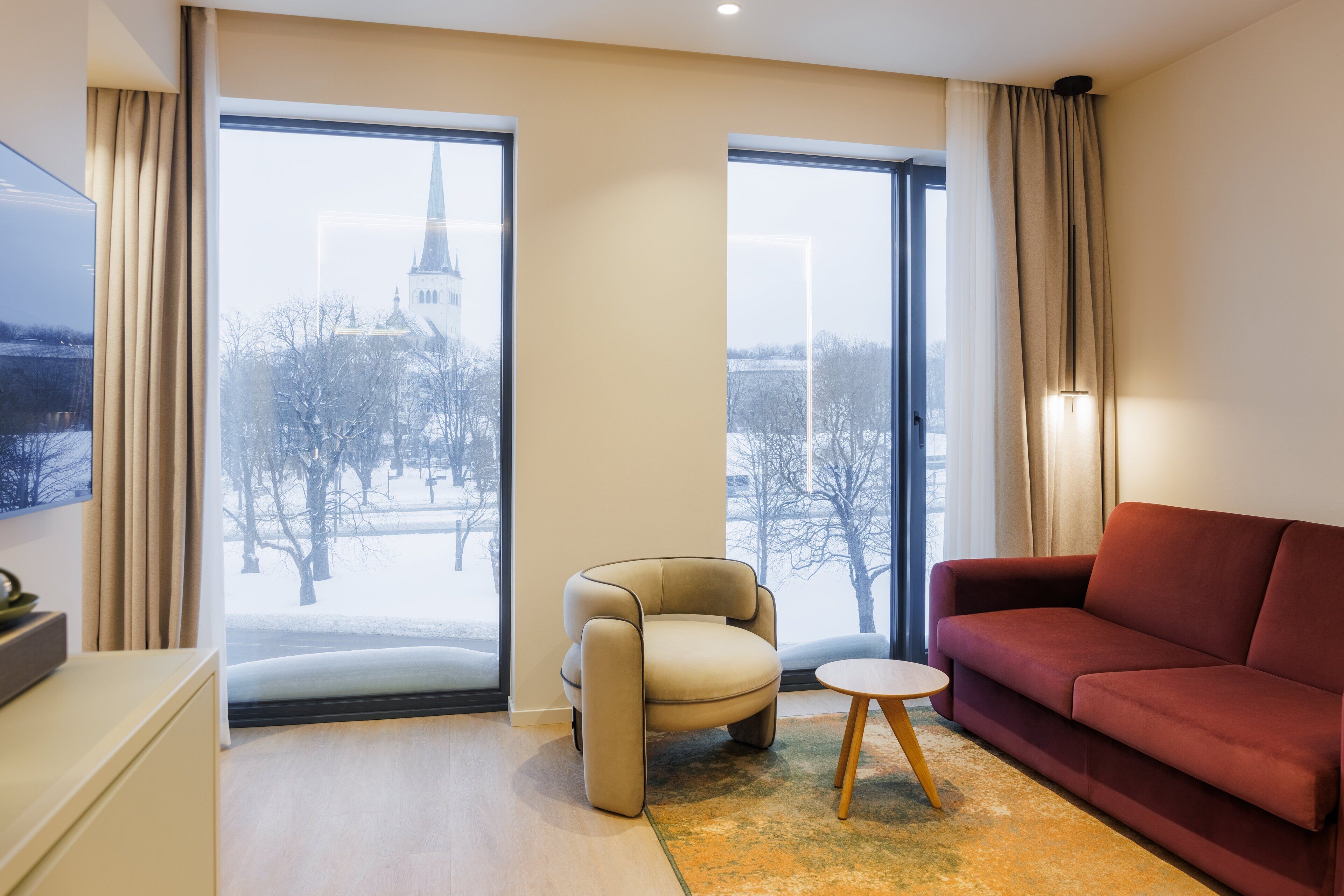 A modern living room with a sofa, armchair, and a small table. Large windows offer a view of a snowy landscape and a church spire.