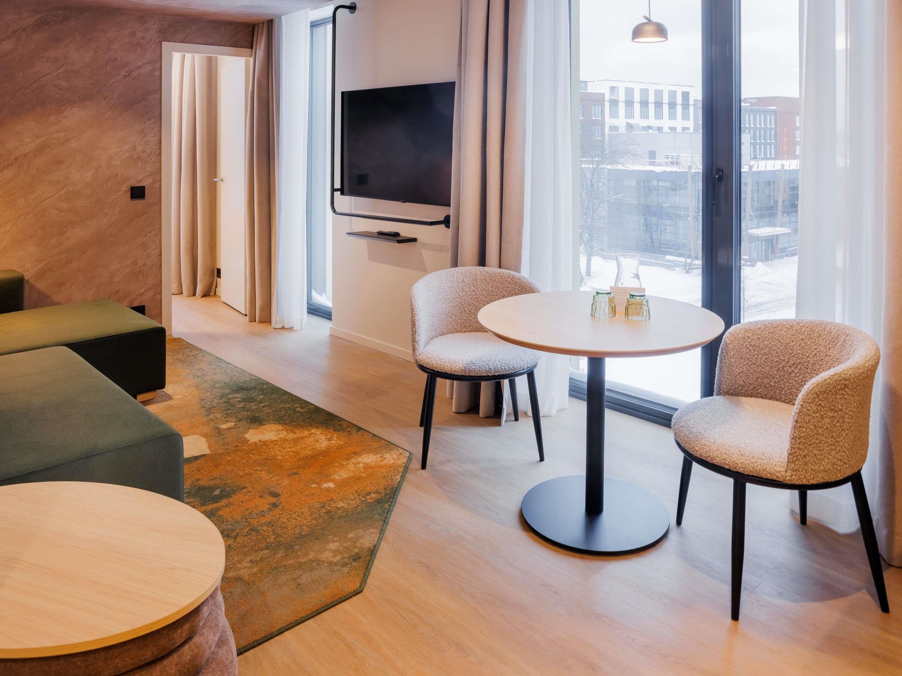A modern living room with a sofa, two armchairs, a round table, a flat-screen TV mounted on the wall, and a large window with a view of buildings.