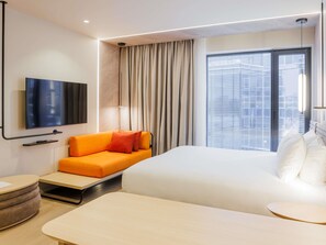 A modern hotel room with a large bed, a sofa, a TV mounted on the wall, and a window with blinds.