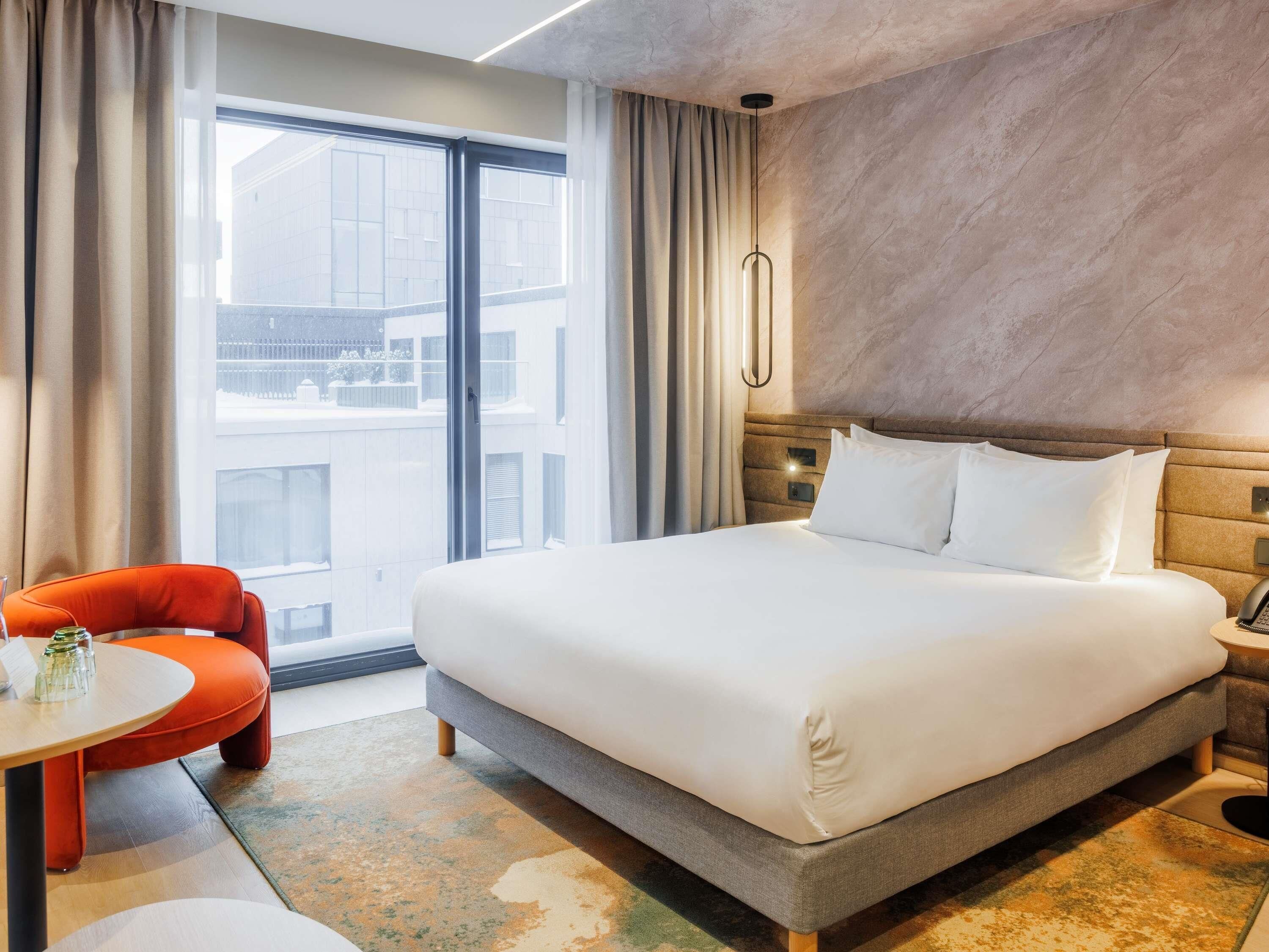 A hotel room with a large bed, a small table with chairs, a telephone, and a view of buildings outside.