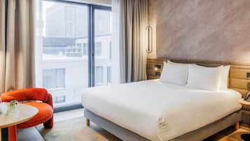 A hotel room with a large bed, a small table with chairs, a telephone, and a view of buildings outside.