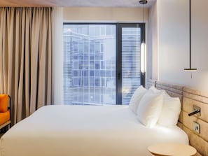 A hotel room with a large bed, a chair, a bedside table, and a view of buildings through the window. - Novotel Tallinn (Tallinn)