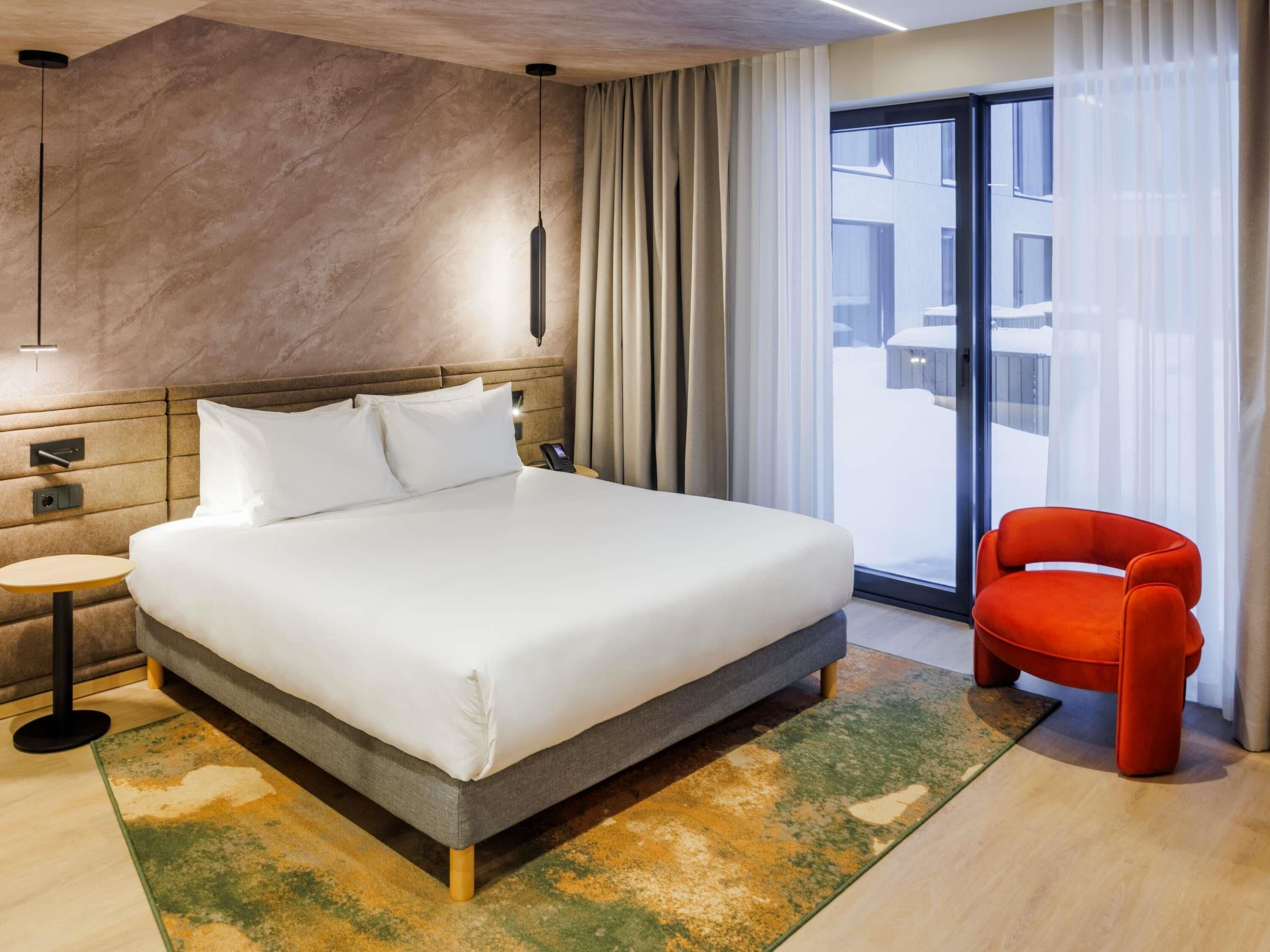 A modern hotel room with a large bed, a small table, a chair, and a view of the outside.