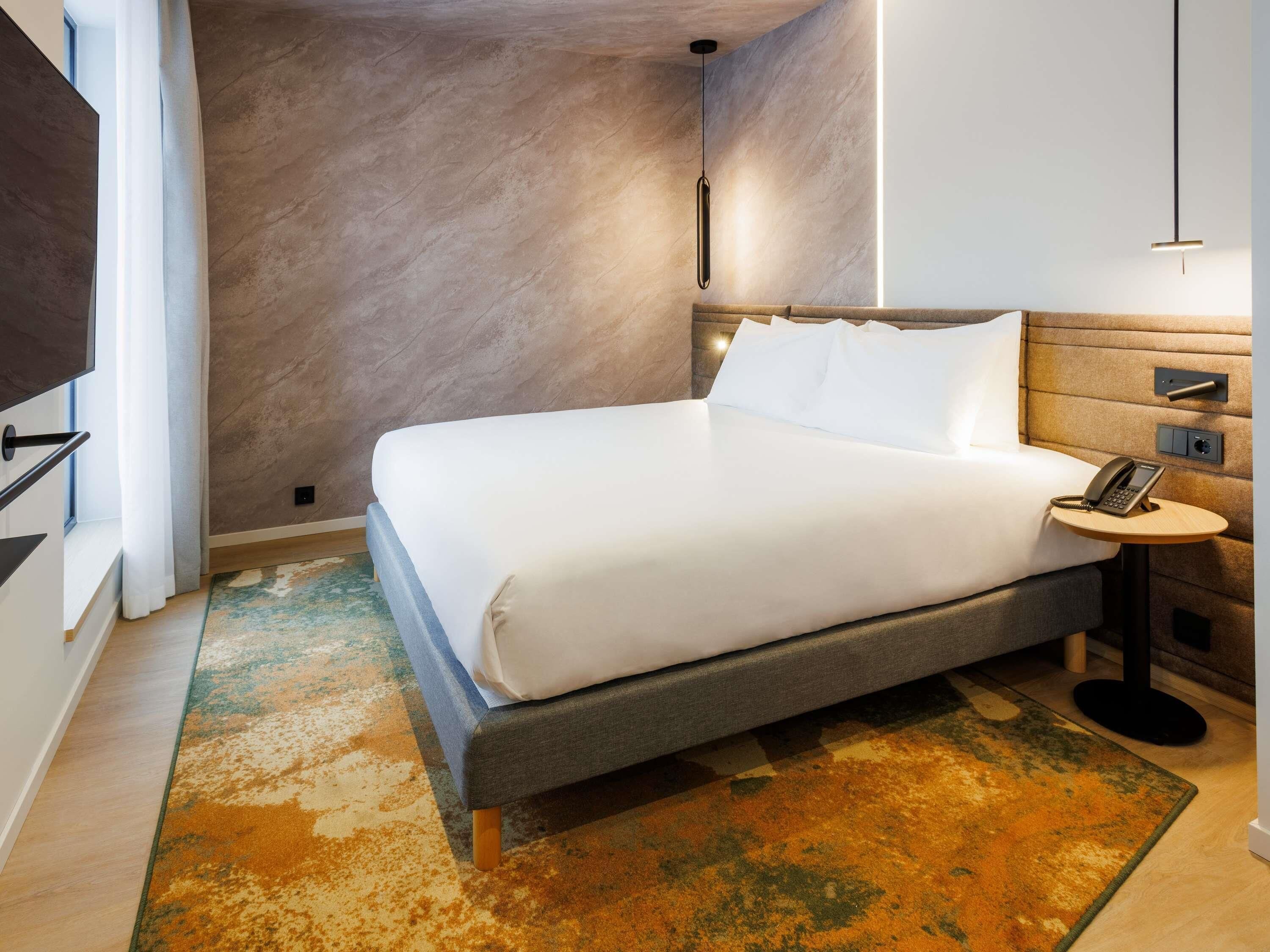 A hotel room with a bed, bedside table with a phone, and a wall-mounted TV.