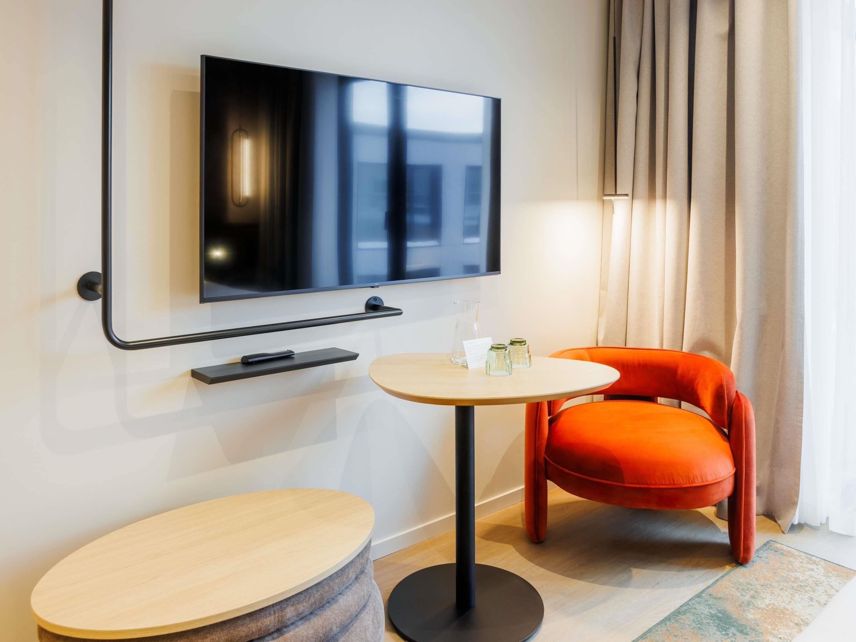 Une chambre d’hôtel moderne équipée d’un téléviseur à écran plat fixé au mur, d’une table ronde en bois, d’une chaise orange et d’une petite table d’appoint avec une lampe et des salières.