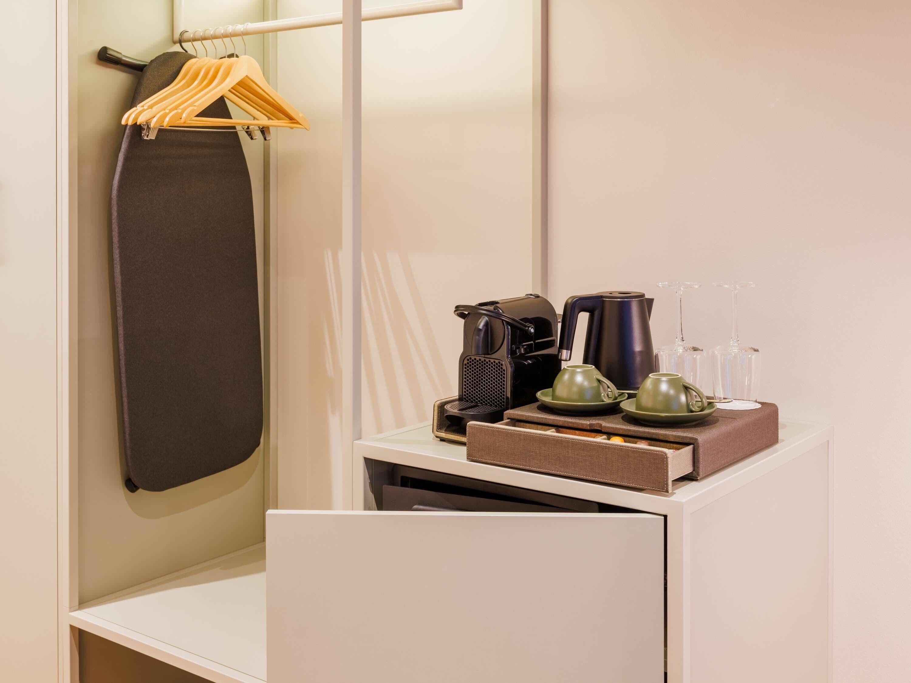 A hotel room with a bed, a hanging garment, a coffee maker, a kettle, and a tray with cups and glasses.