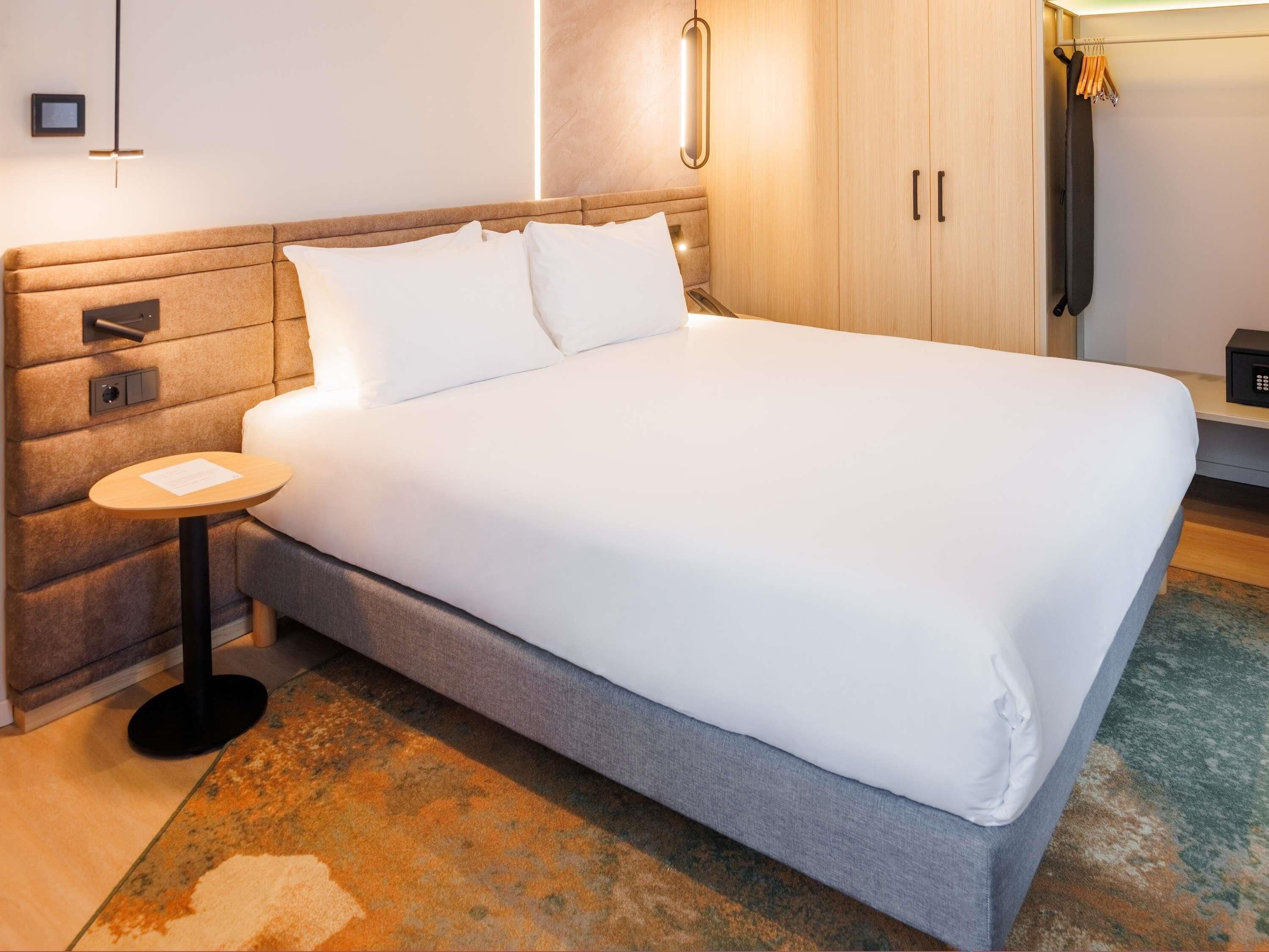 Una cama bien tendida en una habitación de hotel, con cabecera, mesita de noche y lámpara de pared.