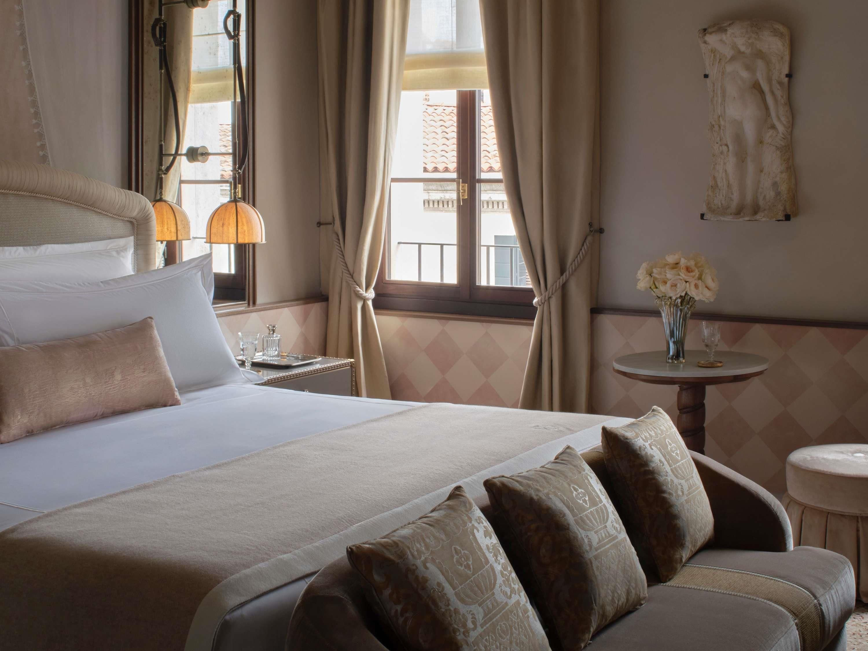 A hotel room with a large bed, a round table with a vase of flowers, and a view of buildings through the window.