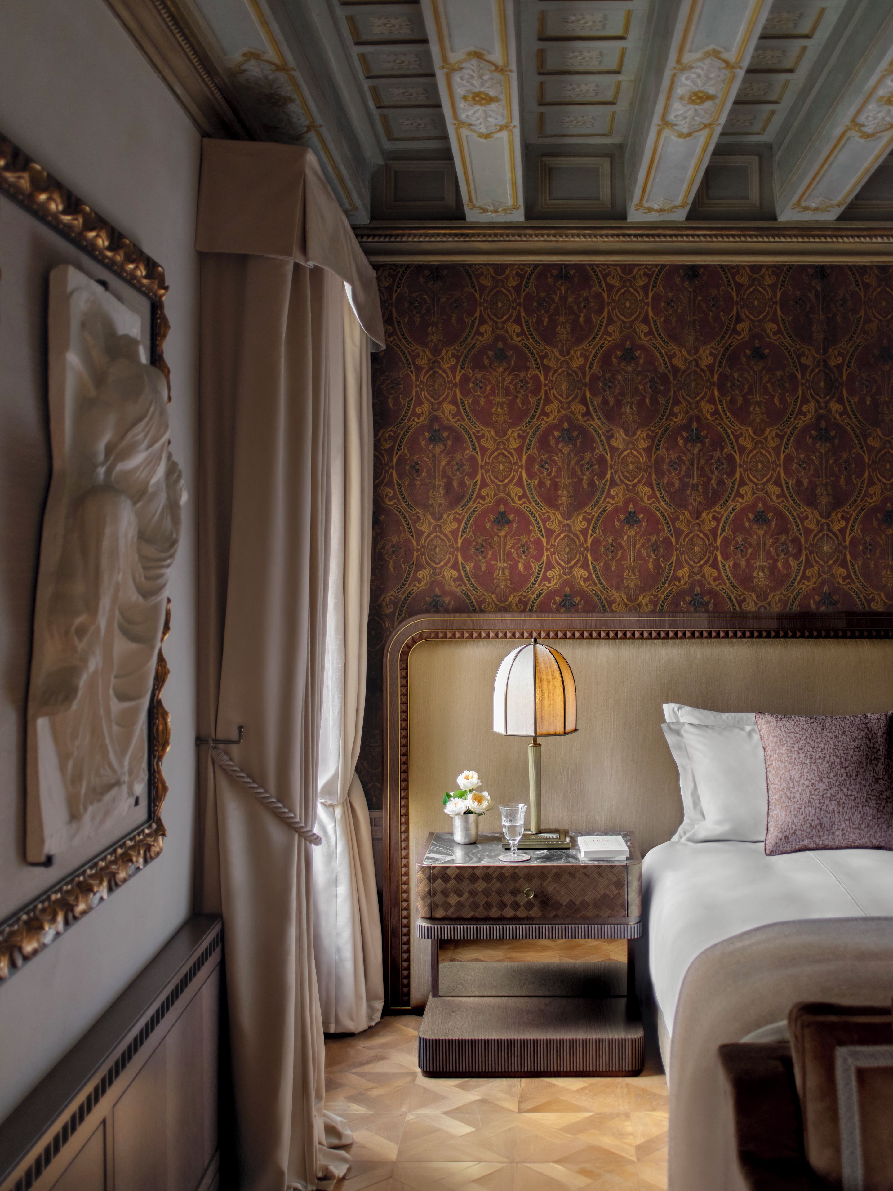 A hotel room with a bed, a nightstand, a lamp, a vase with flowers, and a wall with a patterned wallpaper.