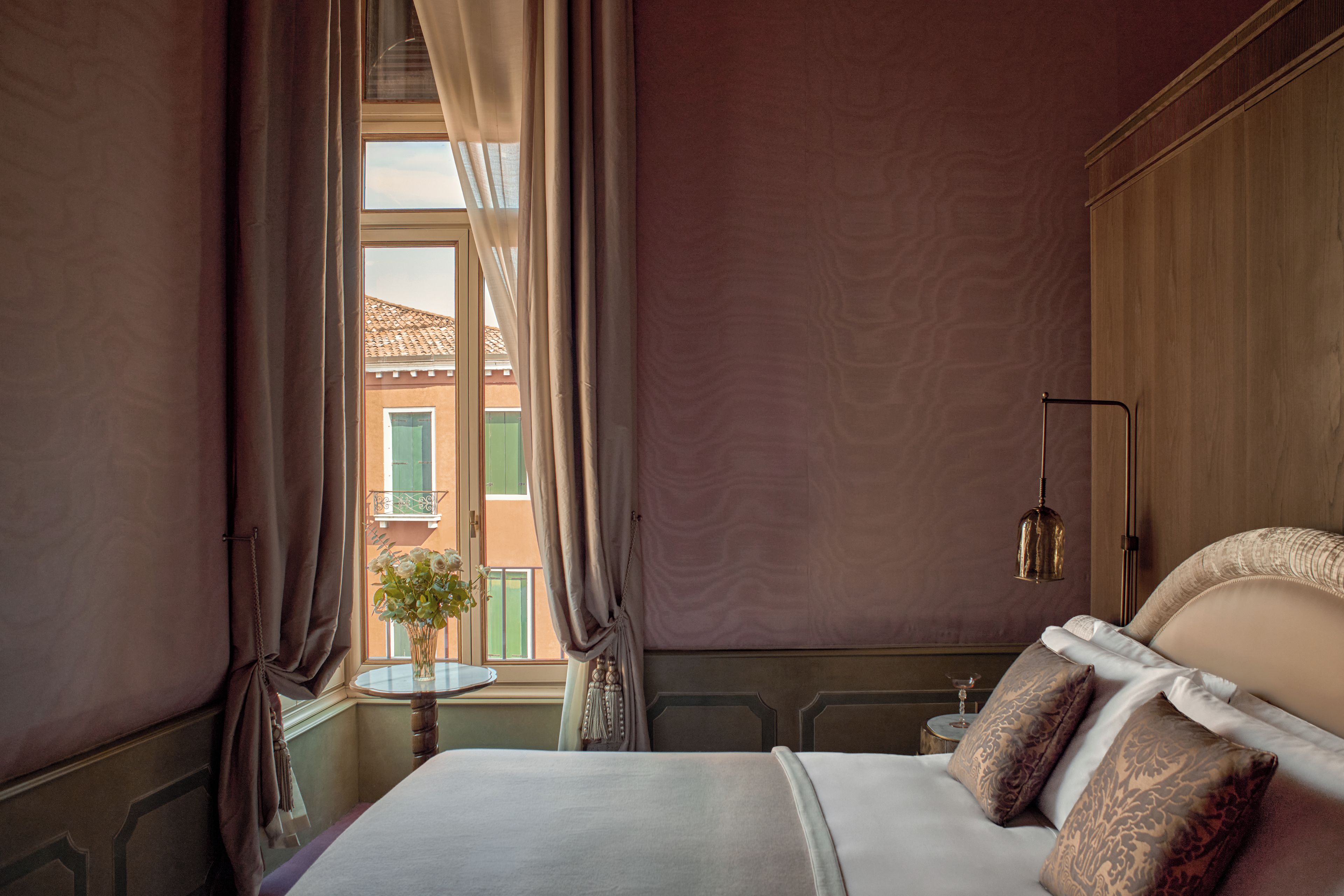 Une chambre d’hôtel avec un lit, une petite table, un vase avec des fleurs, une lampe et une vue sur les bâtiments à l’extérieur de la fenêtre.