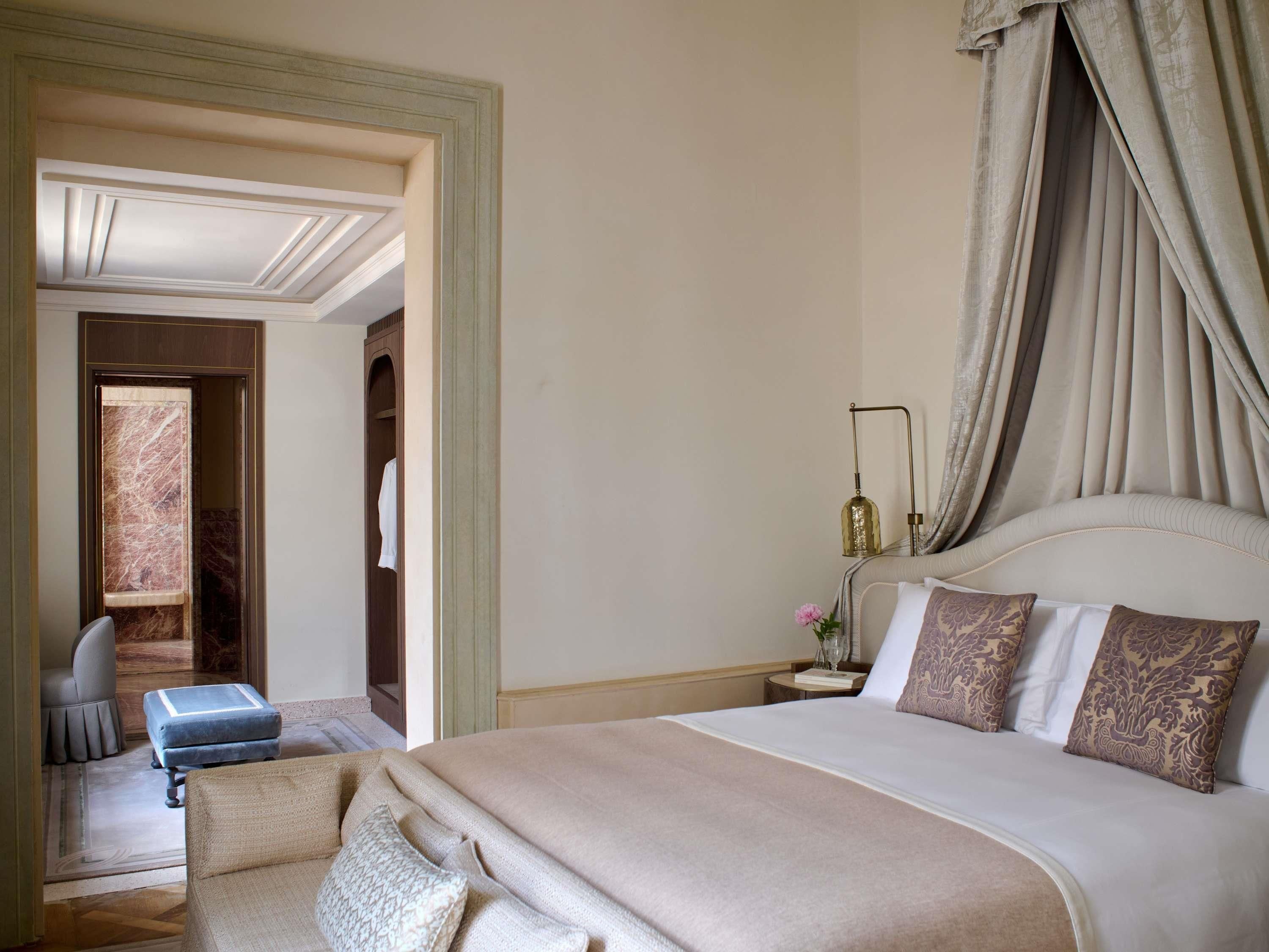 A hotel room with a large bed, a bedside table with a lamp, a chair, and a view of a marble wall.
