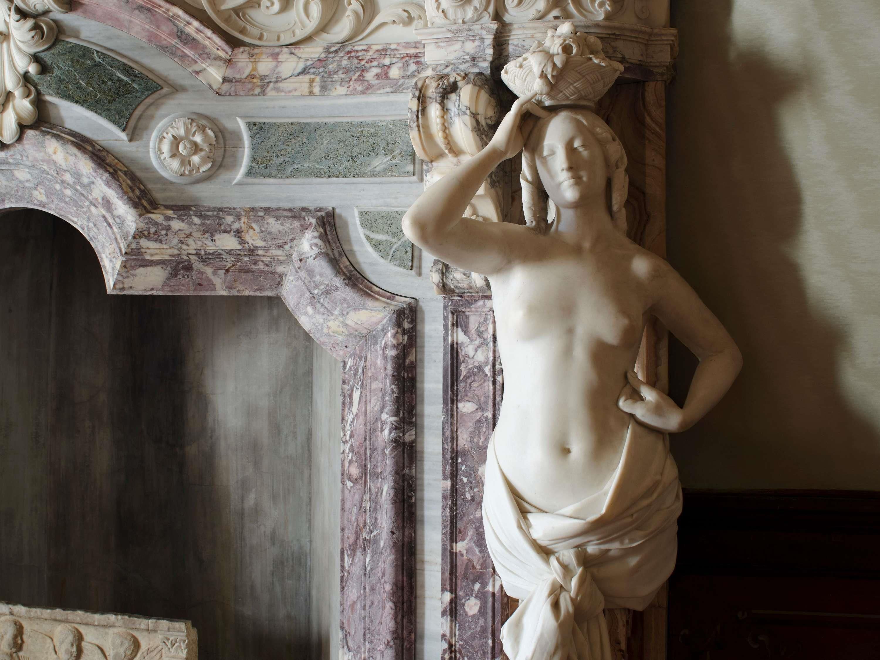 A marble statue with a draped garment, set against a wall with ornate architectural details.