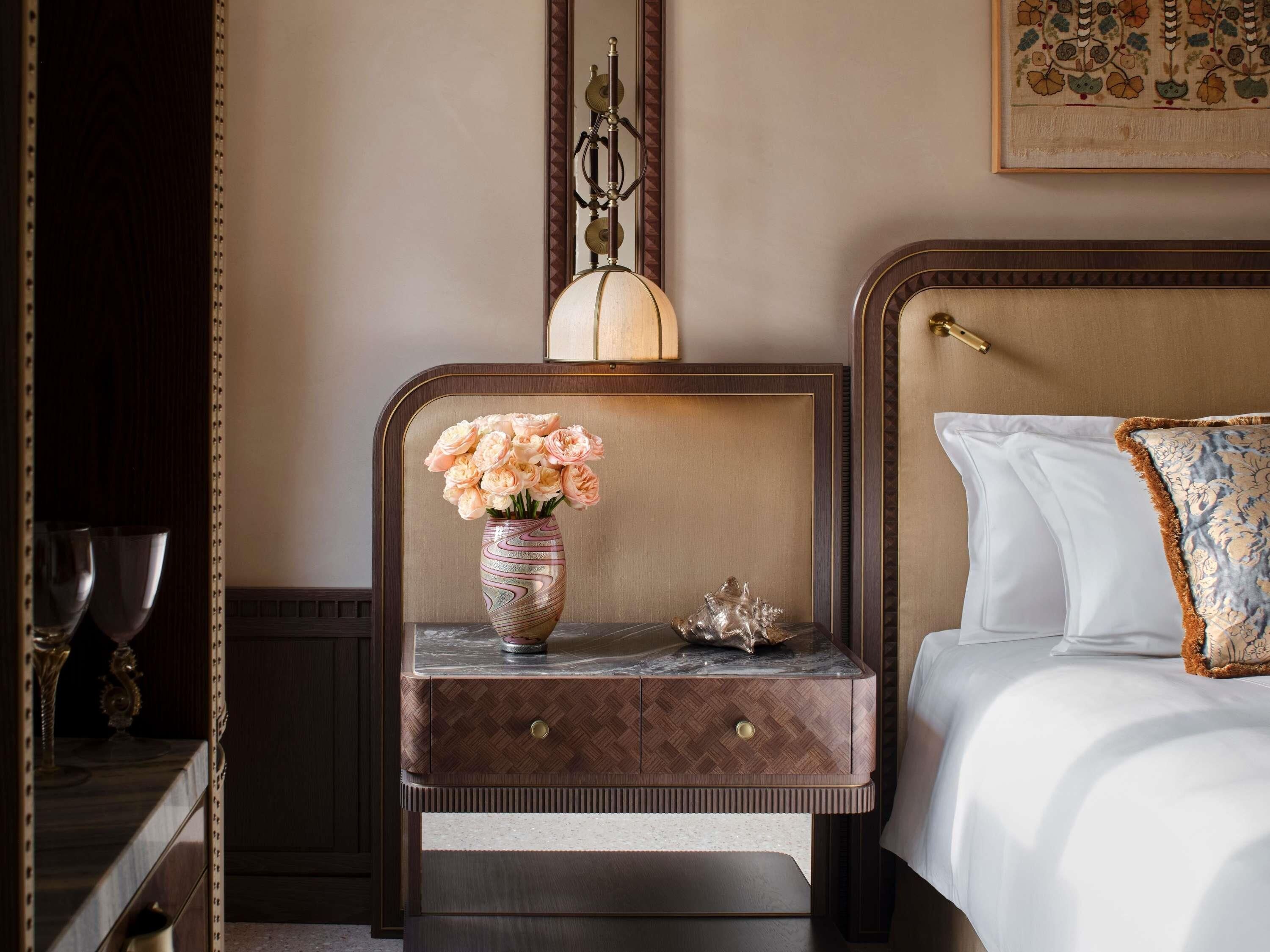 A neatly arranged bedroom with a bed, a nightstand with a vase of flowers, and a wall-mounted lamp.