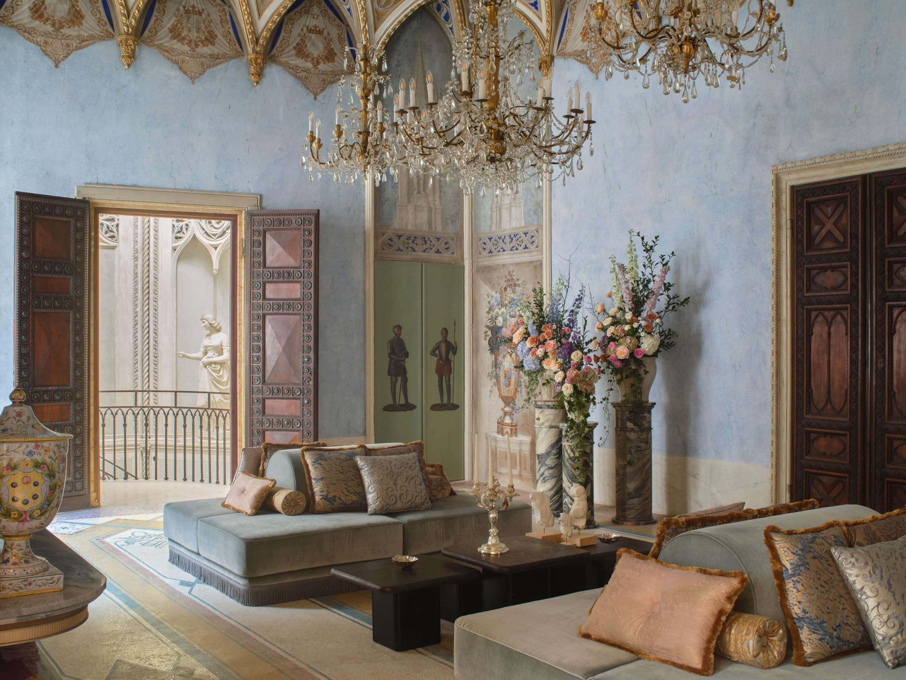 Interior luxuoso com lustres ornamentados, um amplo conjunto de sofás e obras de arte clássicas.