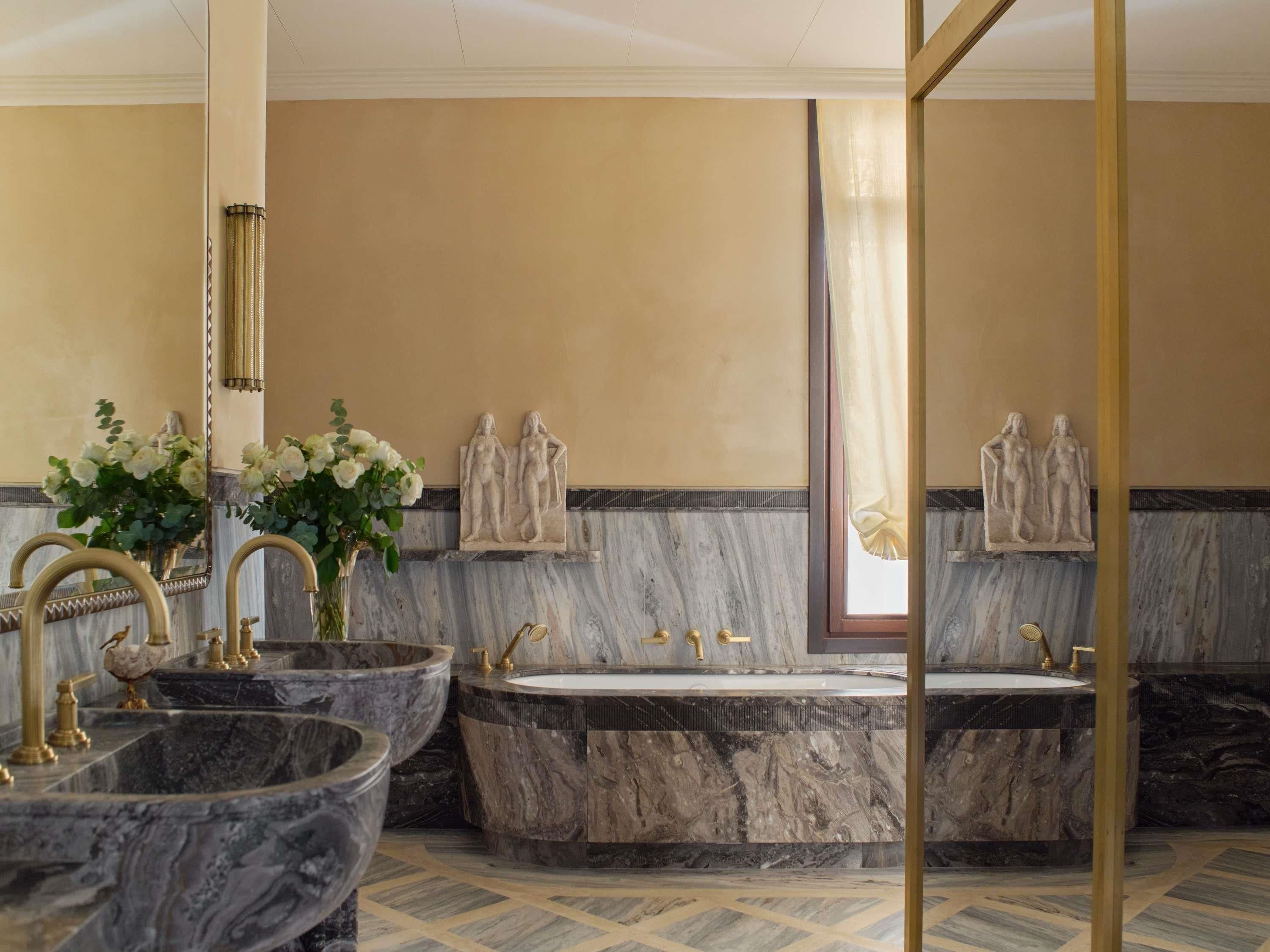 Een luxe badkamer met twee marmeren wastafels, een vrijstaand bad en decoratieve wanddecoraties.